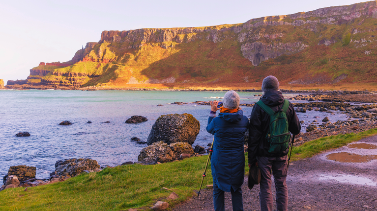 Two visitors admiring the Causeway Cliffs from Port Noffer, with one taking a picture on their phone. Both visitors are wrapped up warm in long coats, hats and scarfs and are both holding hiking sticks.